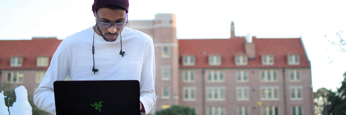 An image of a man sitting outside focused on and looking at his computer with an FSU building in the background.